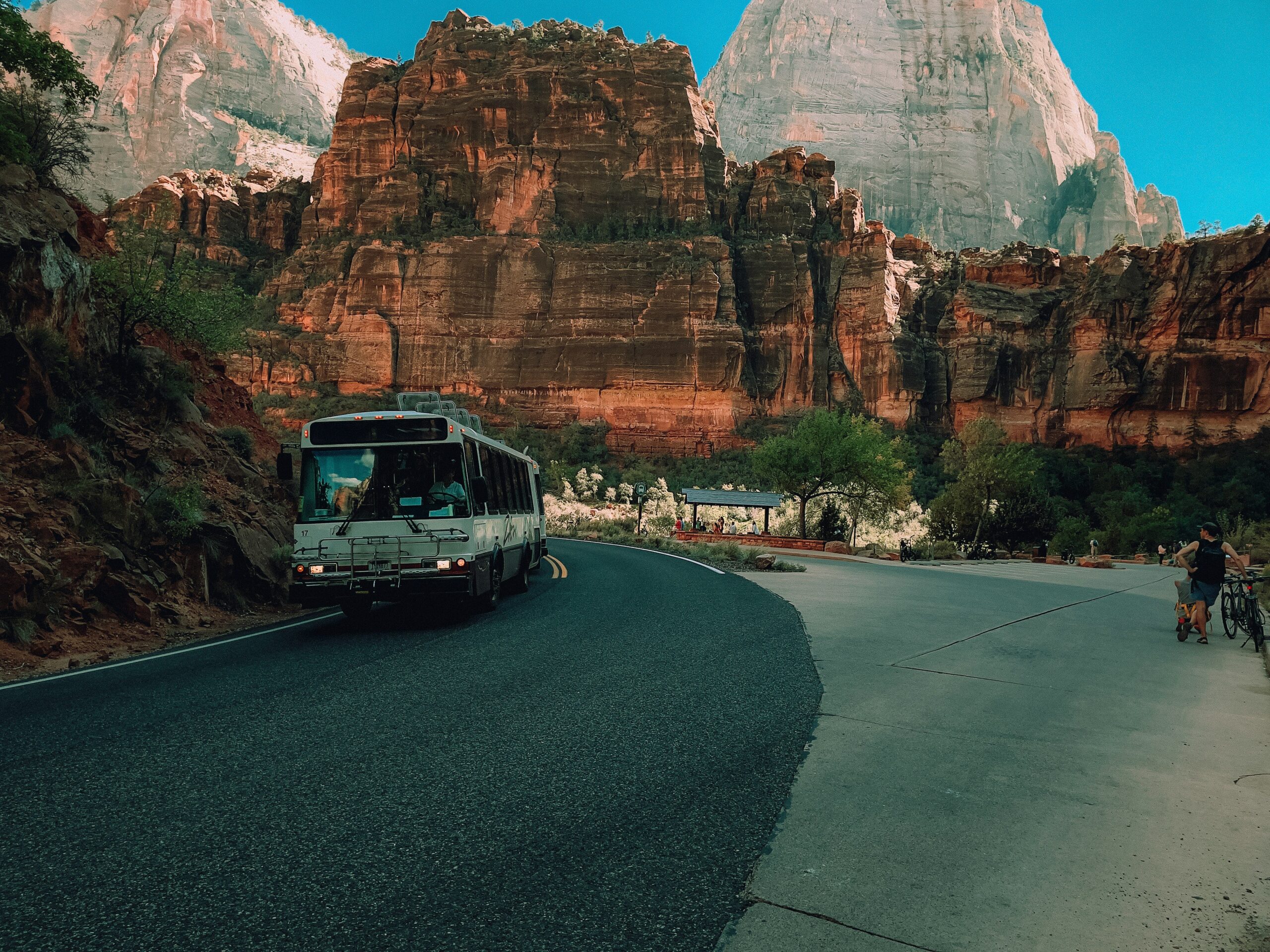 Shuttle bus driving along a winding road through towering red rock cliffs in Zion National Park while visitors walk and cycle nearby