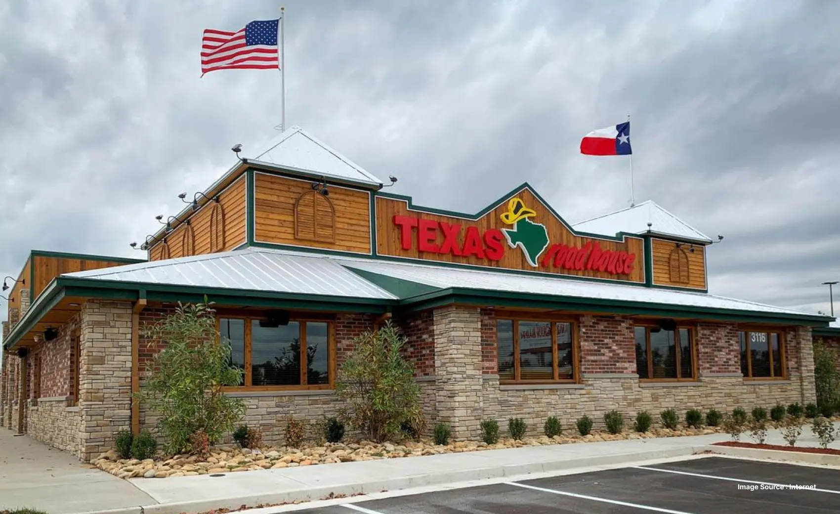 The image shows the exterior of a Texas Roadhouse location. It features the restaurant's signature red and green signage along with the American and Texas flags flying above the building. The design combines rustic wood and stone, with large windows along the side and a well-maintained parking lot.